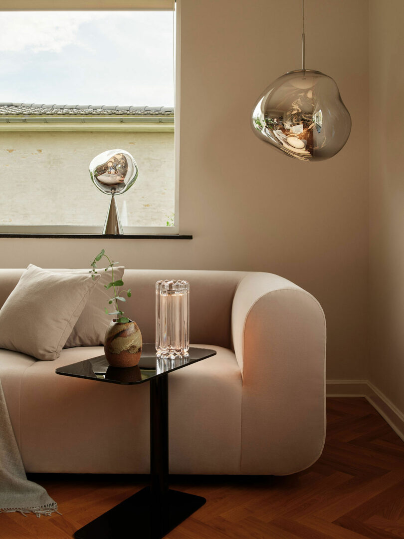 Modern living room with a white sofa, side table holding a glass vase and decorative piece, and two organic-shaped pendant lights; one hanging from the ceiling and one on the windowsill