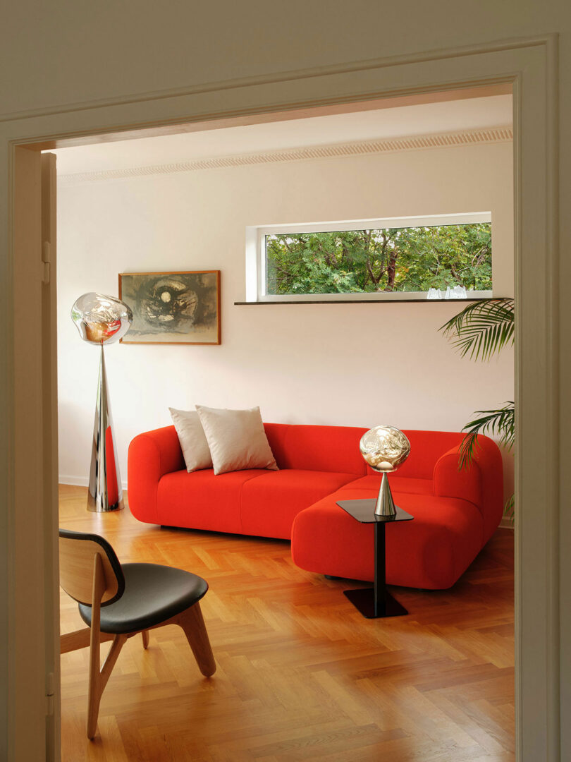 A modern living room with a red sofa, a small black side table, a chair, two floor lamps, and a painting on the wall. A small window offers an outdoor view