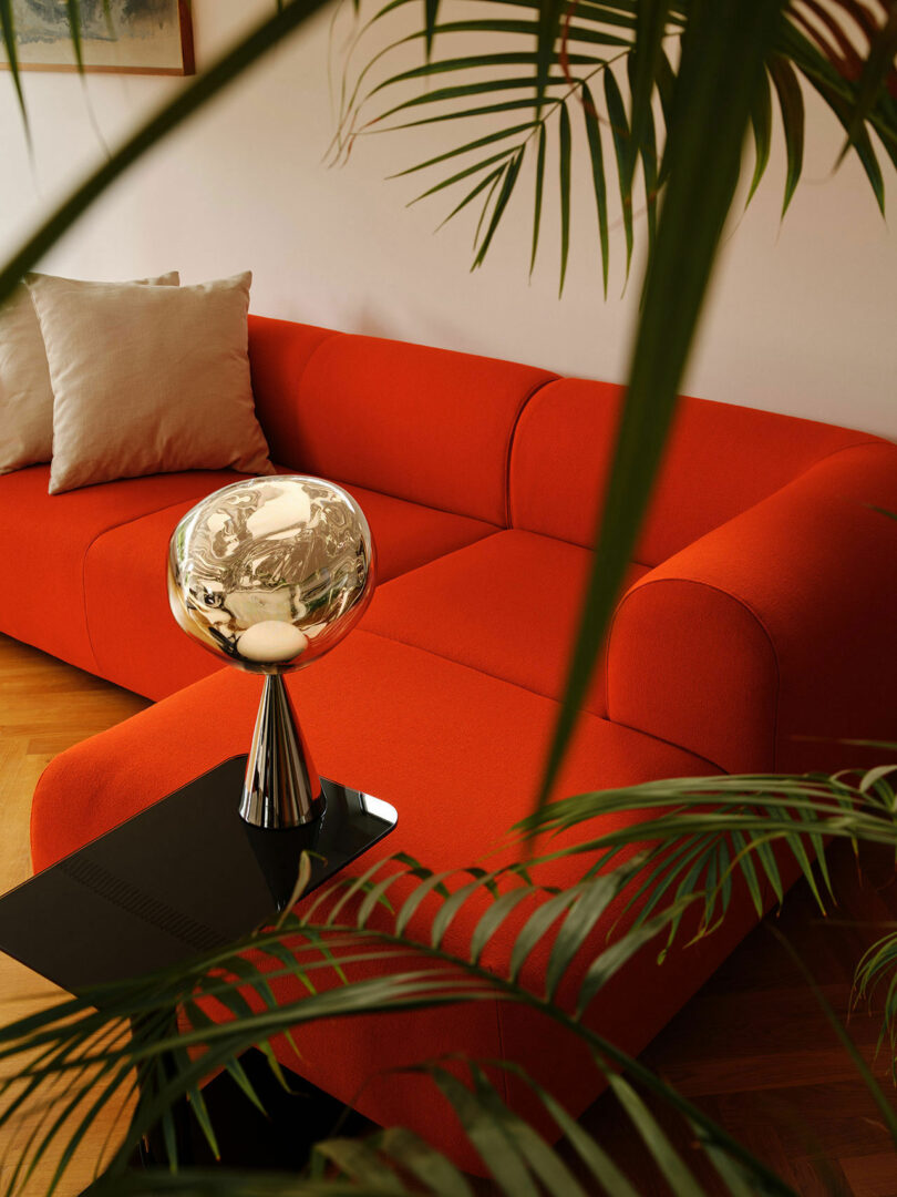 A modern living room features an orange couch with beige pillows, a black side table with a silver globe sculpture, and lush green plants in the foreground