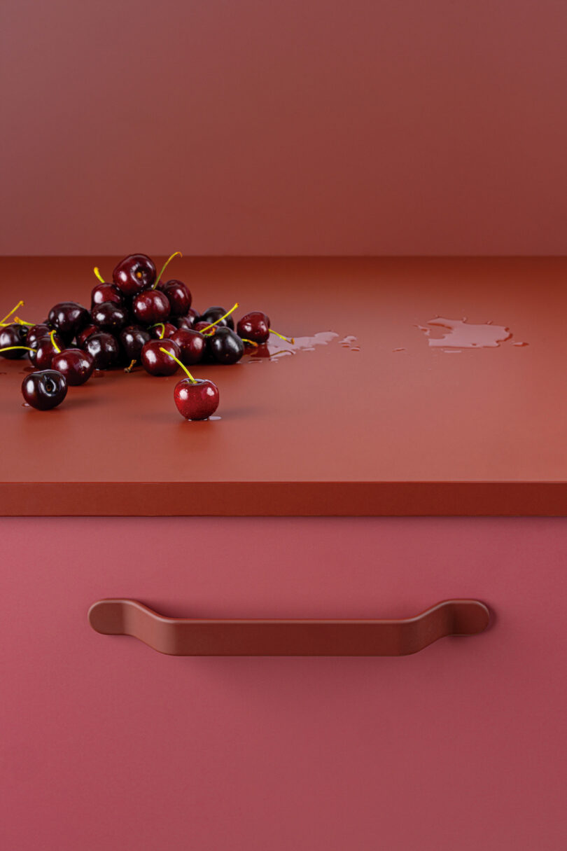 A group of cherries is arranged on a smooth, red surface, with one cherry separated in the foreground. There is a small puddle of water near the cherries