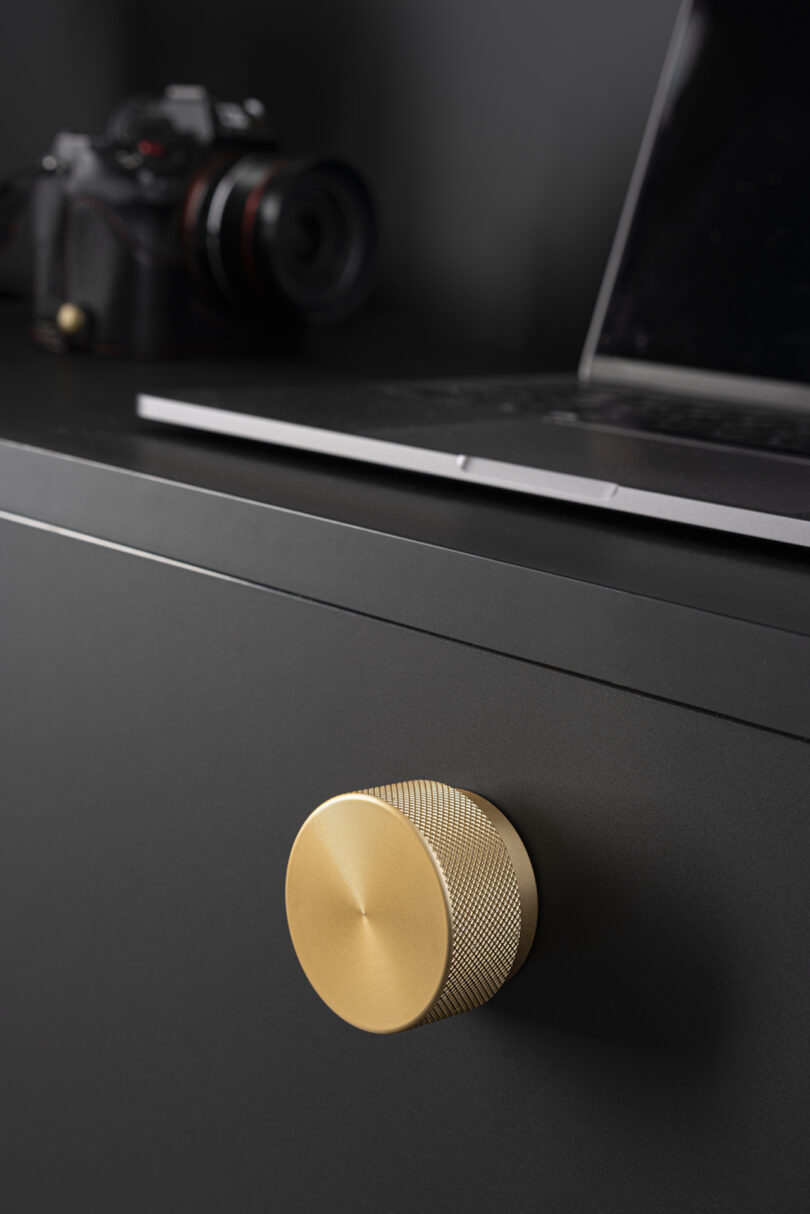 Close-up of a matte black cabinet with a gold round knob. A laptop and a camera are on top of the cabinet