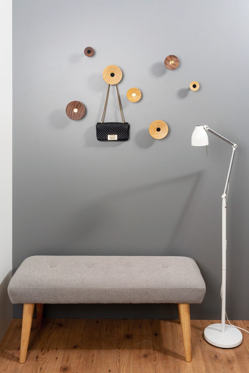 Gray bench with wooden legs against a gray wall with round wooden wall hooks holding a small black handbag. A white floor lamp stands on a wooden floor beside the bench