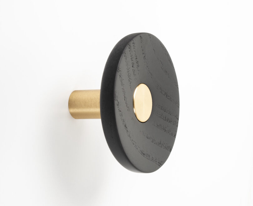 A wall hook with a circular black wooden front and a gold metal body is mounted on a white surface