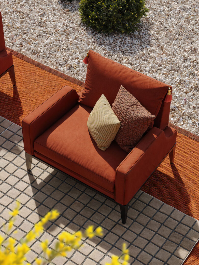 An orange outdoor chair with two patterned pillows is positioned on a tiled patio next to a gravel area, bordered by an orange rug