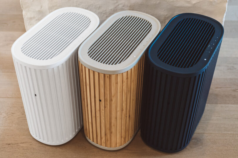 Three air purifiers in different colors—white, wood-grain, and black—are placed side by side on a wooden floor.