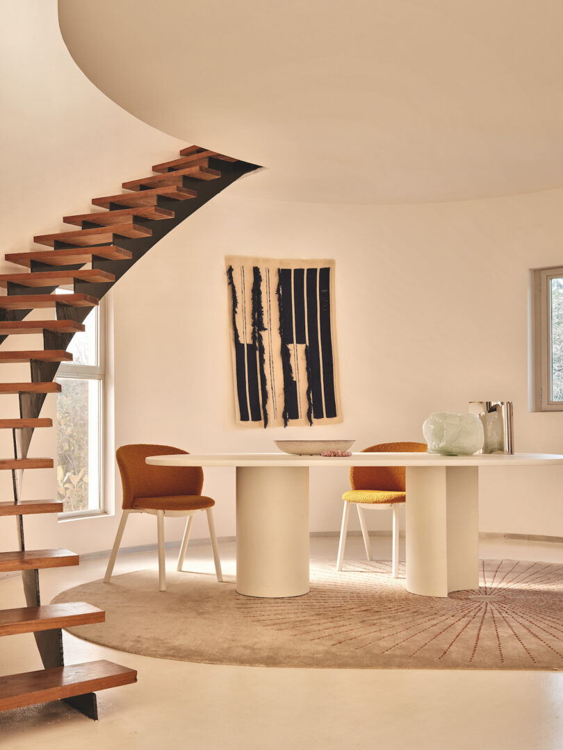 A modern interior features a wooden spiral staircase, two white cylindrical tables, two mustard chairs, a beige rug, and abstract wall art. The room has minimalistic decor and natural light