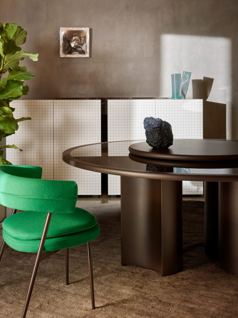 A modern dining area with a round table, a green chair, a white checkered cabinet in the background, and a potted plant on the left. A sculpture and glass items are on the table and cabinet