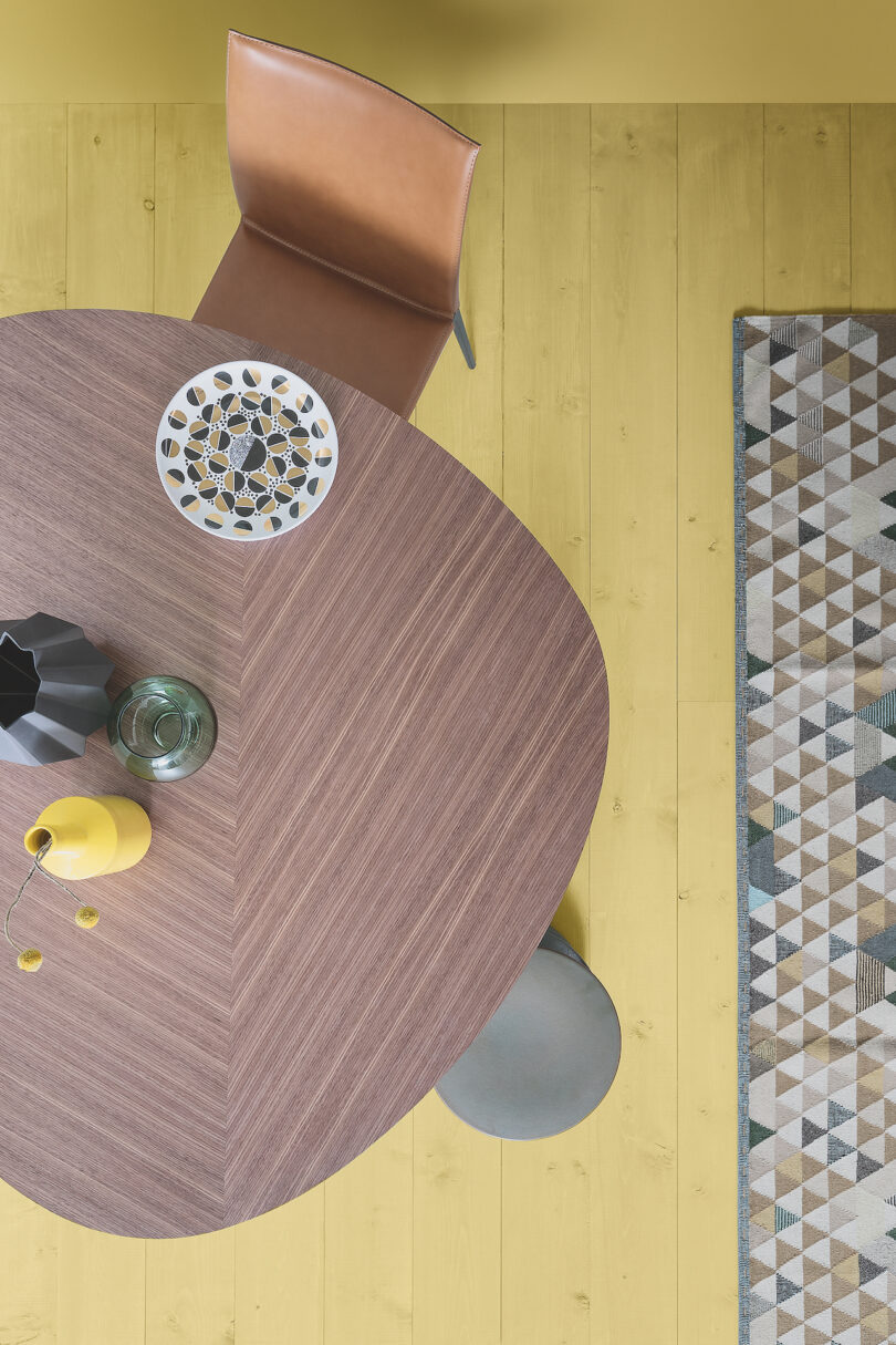 Aerial view of a wooden dining table with a geometric-pattern dish, vase, and drink. A leather chair and patterned rug are on a light-colored wooden floor