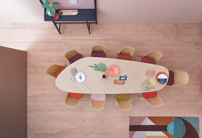 Aerial view of a dining table set for eight with mismatched chairs, plates, and decorative items. A sideboard with plants and decor is positioned along the wall. The room has wooden flooring and a rug