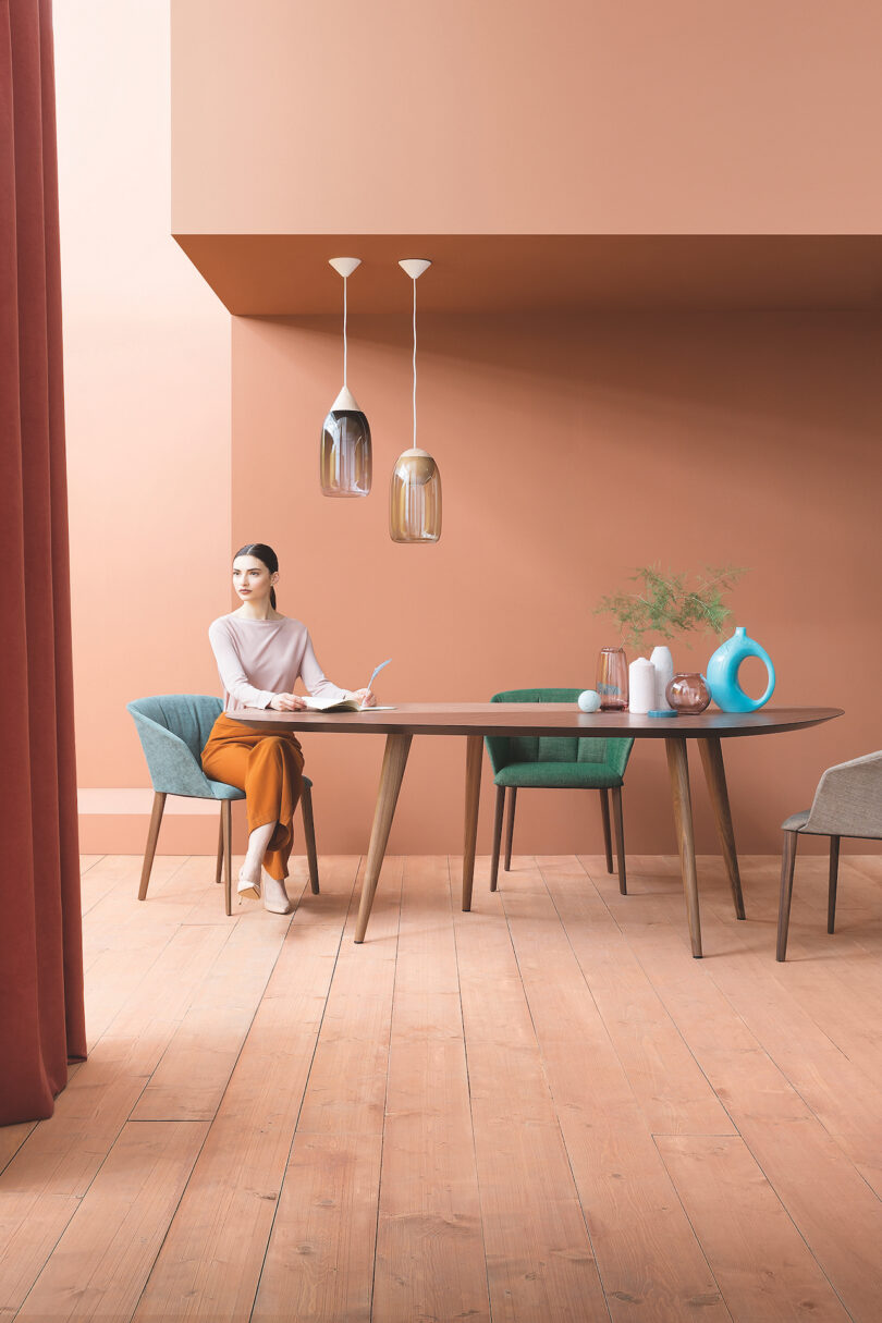 A person sits at a modern wooden dining table with various decorative items, against a peach-colored wall. The room features large pendant lights, chairs in different colors, and wooden flooring