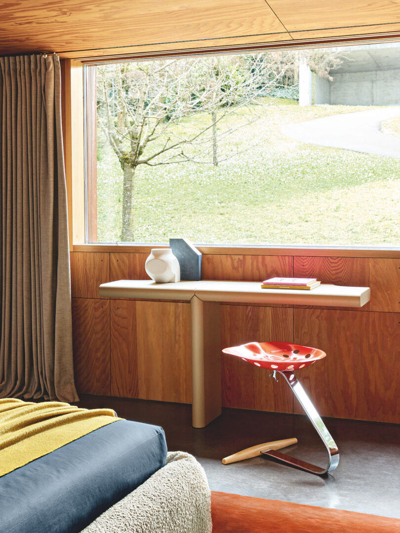 A room with a large window showing a grassy landscape outside, furnished with a light wood desk, red chair, and decor items including vases and books. A bed with mustard and navy bedding is visible