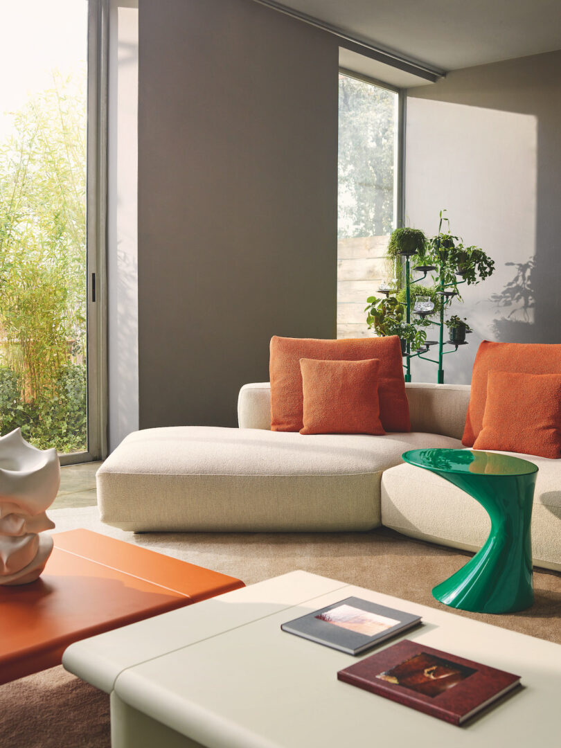 A modern living room featuring a white sofa with orange cushions, a green side table, orange coffee table, books, and a decorative white sculpture. Large windows allow ample natural light to enter