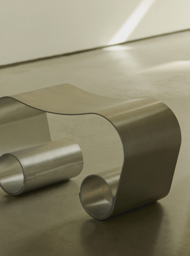 A metallic bench with a wavy, modern design sits on a smooth floor in a minimalist setting.