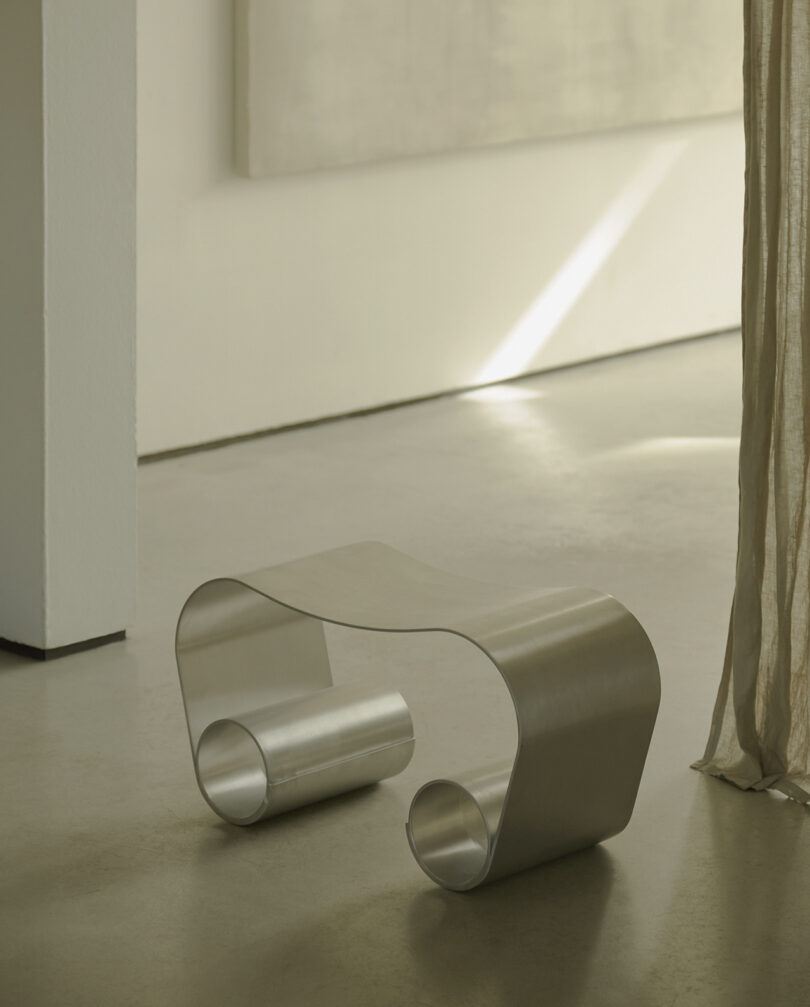 A sleek, metallic, curvy bench sits on a smooth, light-colored floor near a translucent curtain. A large, plain canvas is on the wall in the softly lit room.