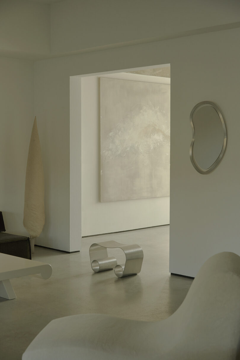 Minimalist room with modern furniture, including a sculptural chair, curved metal table, abstract wall art, and a decorative metal wall piece on a neutral-colored wall.