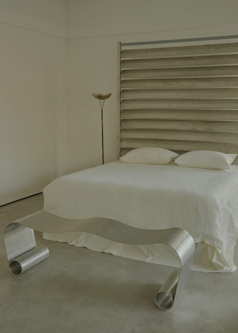 Minimalist bedroom with a white bed, metallic wave-shaped bench, floor lamp, and art piece with horizontal slats on the wall.