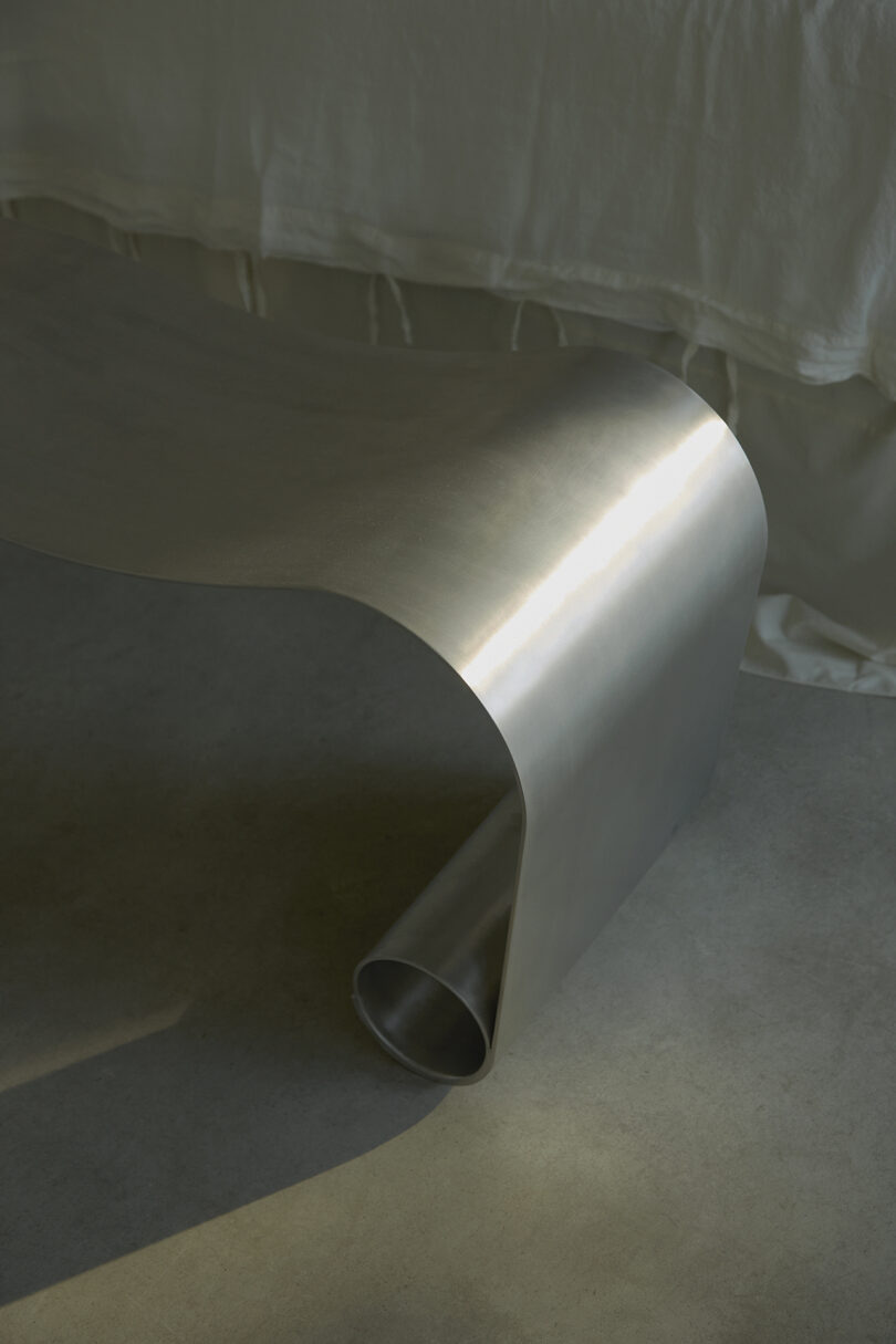 A sleek, metallic bench with a curved design sits on a concrete floor near a white fabric.