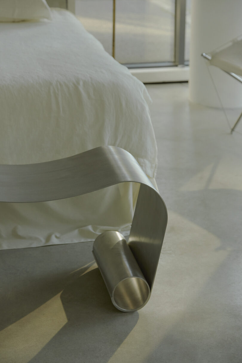 Modern minimalist bedroom with a curved metal bench at the foot of a bed with white linens, next to a window with light streaming in.