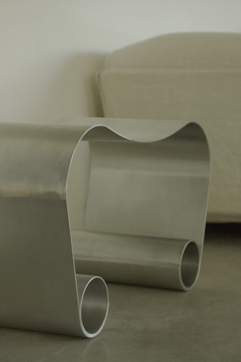 A modern, silver, metal coffee table with a smooth, curved design. A beige couch is in the background.