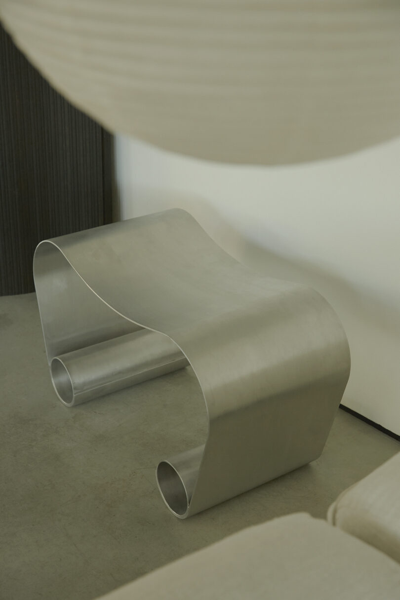 A sleek, metallic, ribbon-shaped chair with a smooth, curved design stands on a concrete floor against a plain wall.