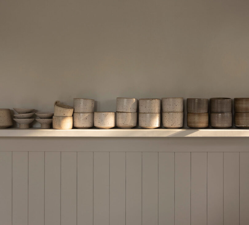 A row of minimalist ceramic pots and bowls in neutral tones are displayed on a shelf against a light-colored wall.