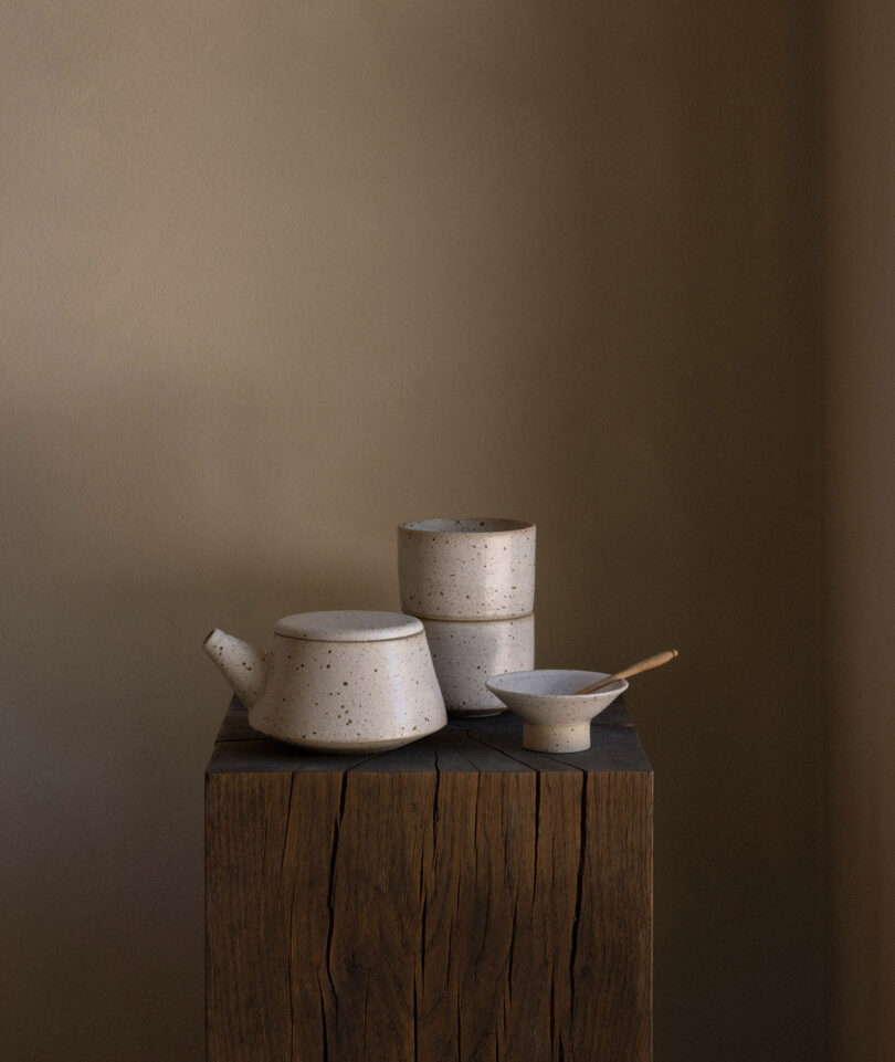 A ceramic teapot, two cups, a bowl, and a spoon sit on a wooden block against a plain brown background.