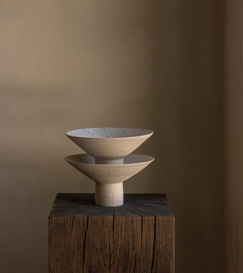 Two stacked, speckled ceramic bowls rest on a wooden pedestal against a neutral background.