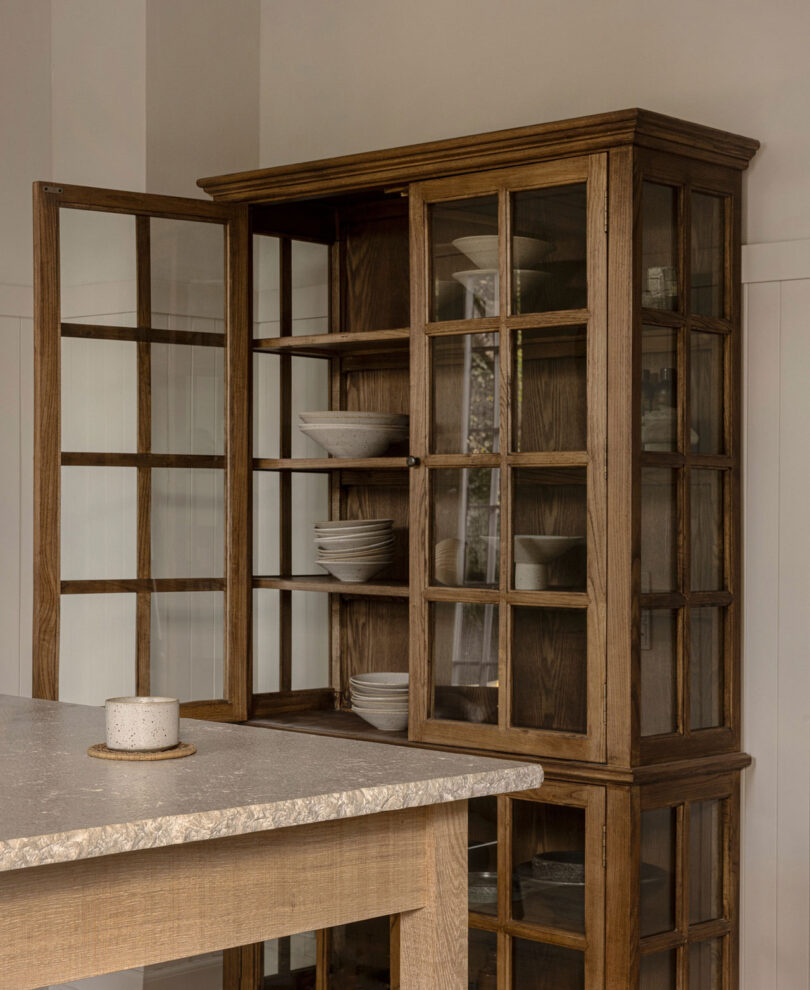 A wooden cabinet with glass doors is open, revealing neatly stacked white dishes. A stoneware cup sits on the countertop in the foreground.