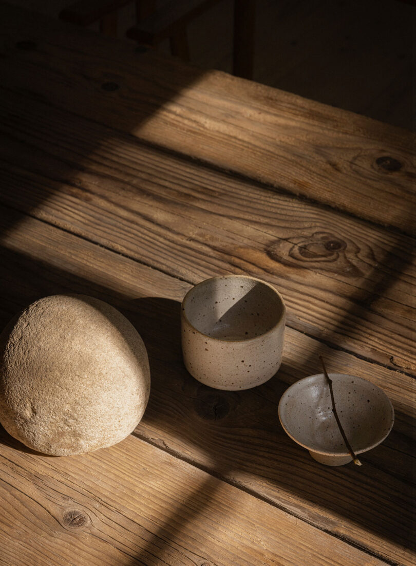 Three ceramic items, including a rounded object, a cup, and a small bowl with a stick, are placed on a wooden table with natural light casting shadows.