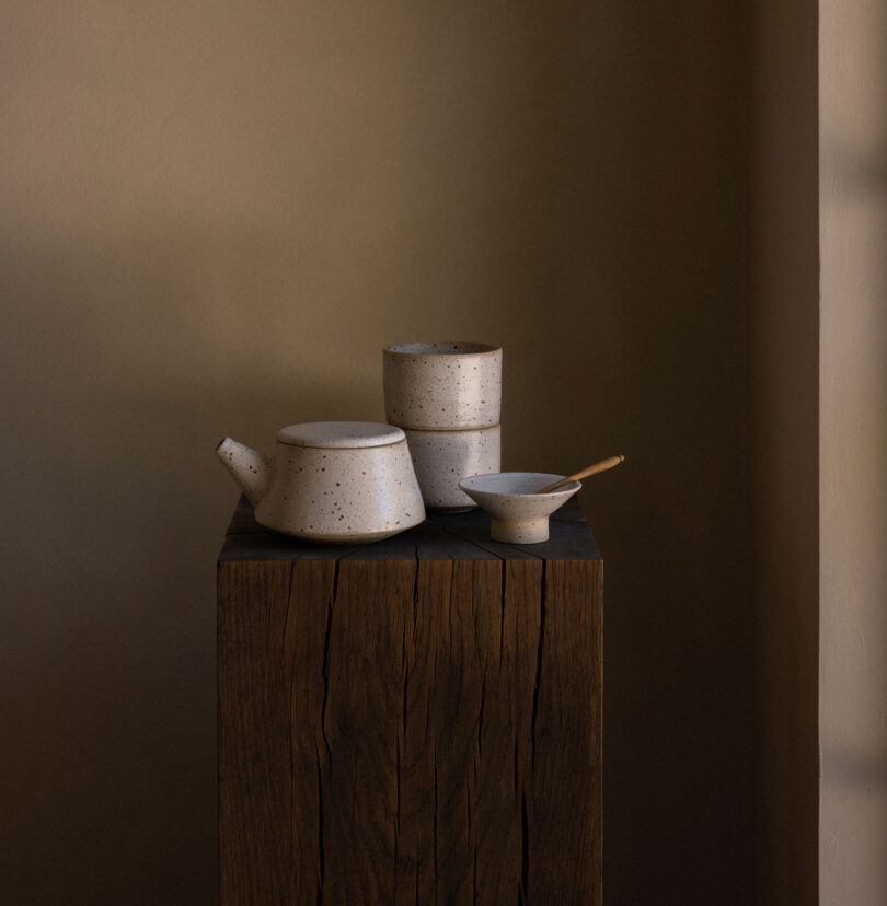 Ceramic teapot, two cups, and a small bowl with a spoon on a wooden pedestal against a neutral background.