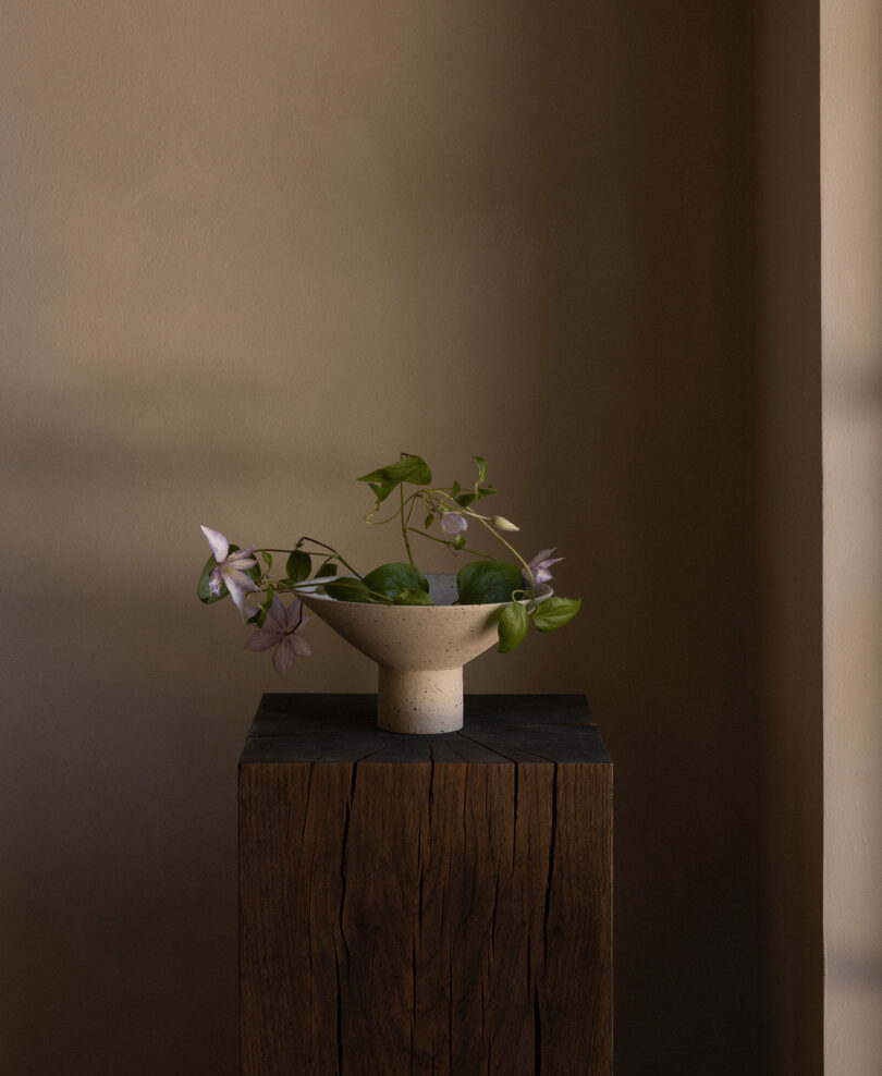 A small ceramic bowl with green and purple flowers is placed on a wooden pedestal against a neutral wall.