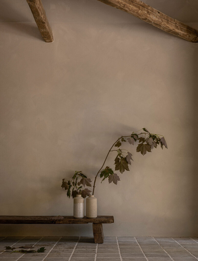 An indoor scene with a rough wooden bench, two ceramic vases containing leafy branches, and exposed wooden beams on the ceiling.