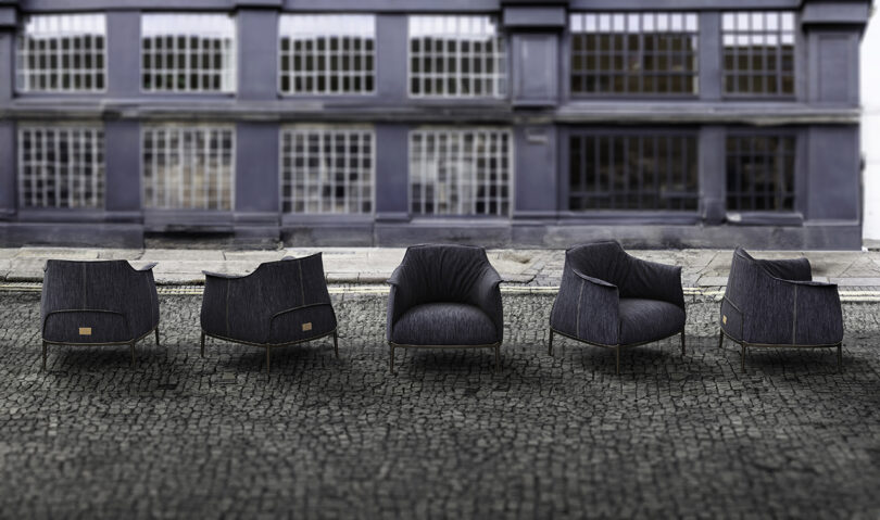 Five modern black armchairs aligned on a cobblestone pavement with a large industrial-style windowed building in the background.