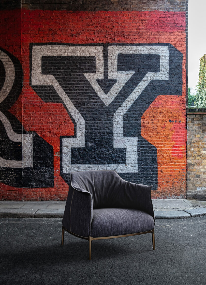 A dark gray armchair is placed on a street in front of a brick wall with large, bold graffiti letters in black, red, and white.