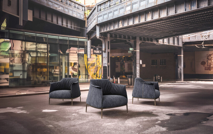 Three modern, dark blue armchairs are arranged on a city street under an elevated train track in an urban setting.