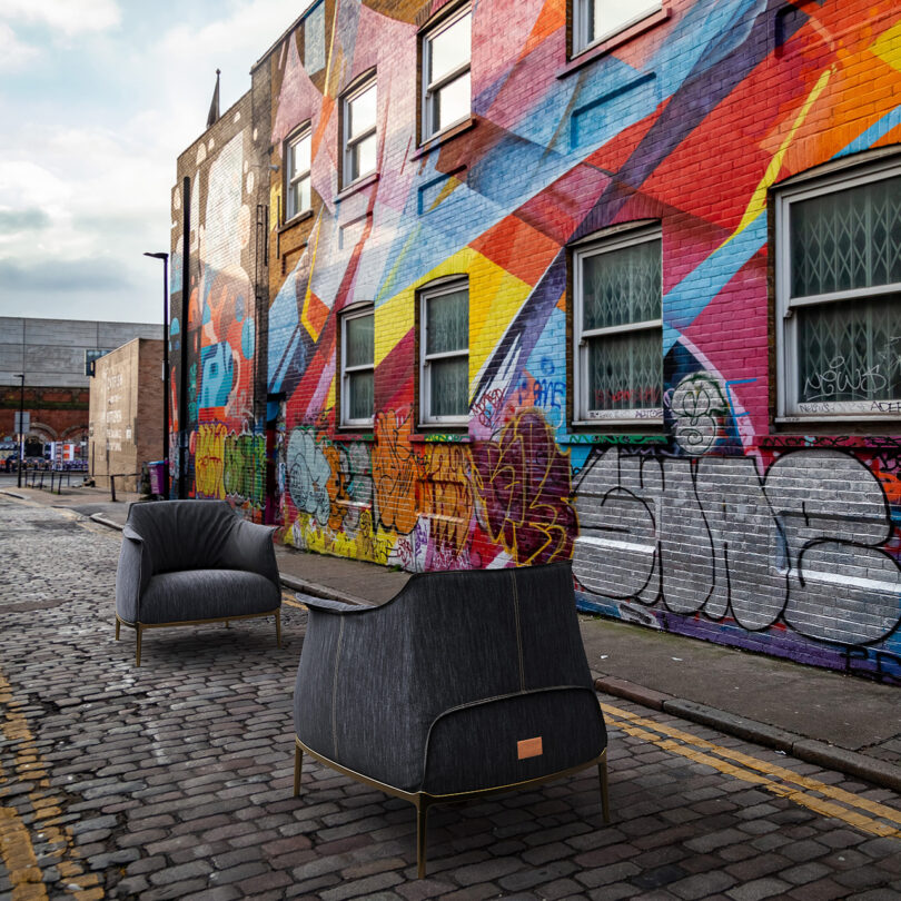 Two chairs on a cobblestone street in front of a colorful, graffiti-covered brick wall with windows.