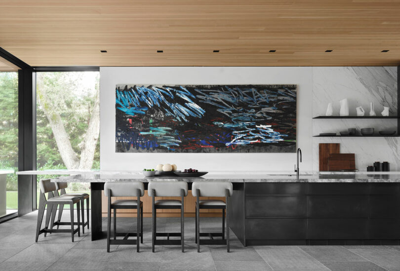 Modern kitchen with a large marble island, barstools, dark cabinetry, white marble backsplash, and a large abstract painting on the wall.