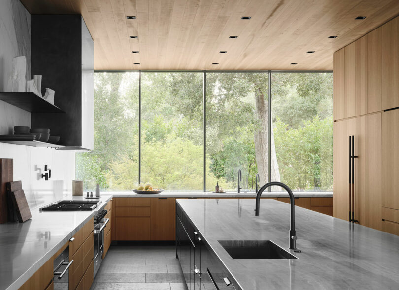 Modern kitchen with a large marble island, under-counter sink, wooden cabinets, and a wall of floor-to-ceiling windows offering views of lush greenery outside.