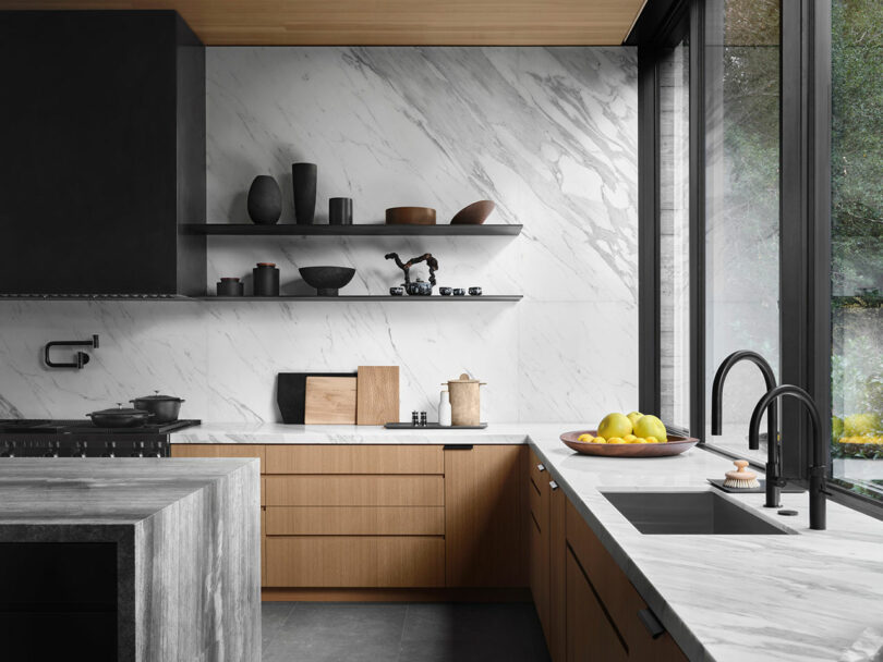 Modern kitchen with marble countertops, wooden cabinets, black fixtures, and a large window. There are shelves with decorative items and a bowl of lemons on the counter.