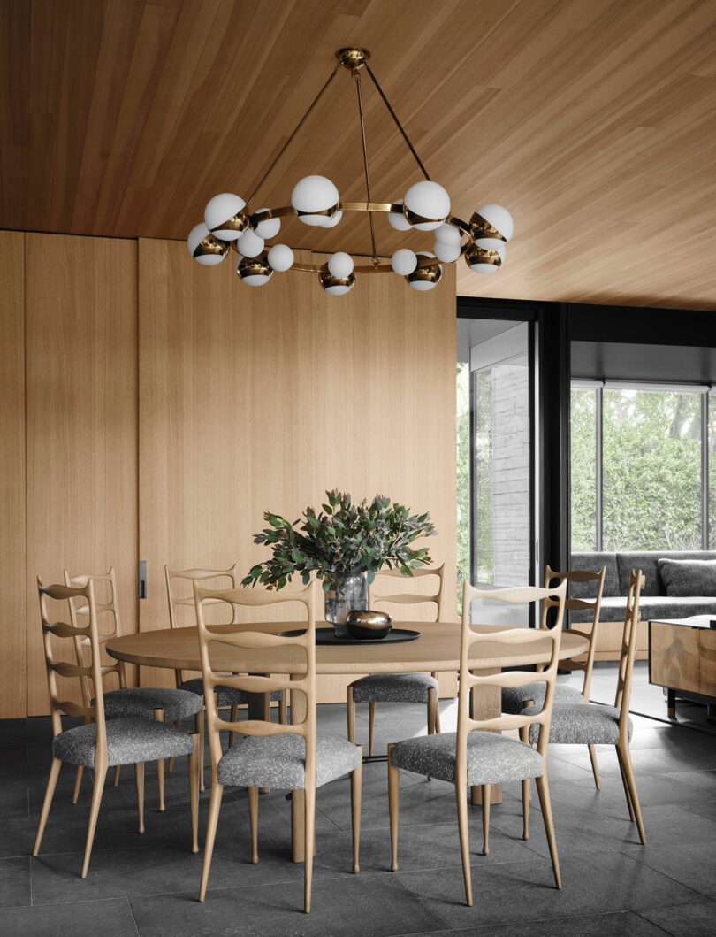 A dining table with eight wooden chairs and a vase of green leaves. A modern chandelier hangs above. The room features wood panels and large windows showing greenery outside.