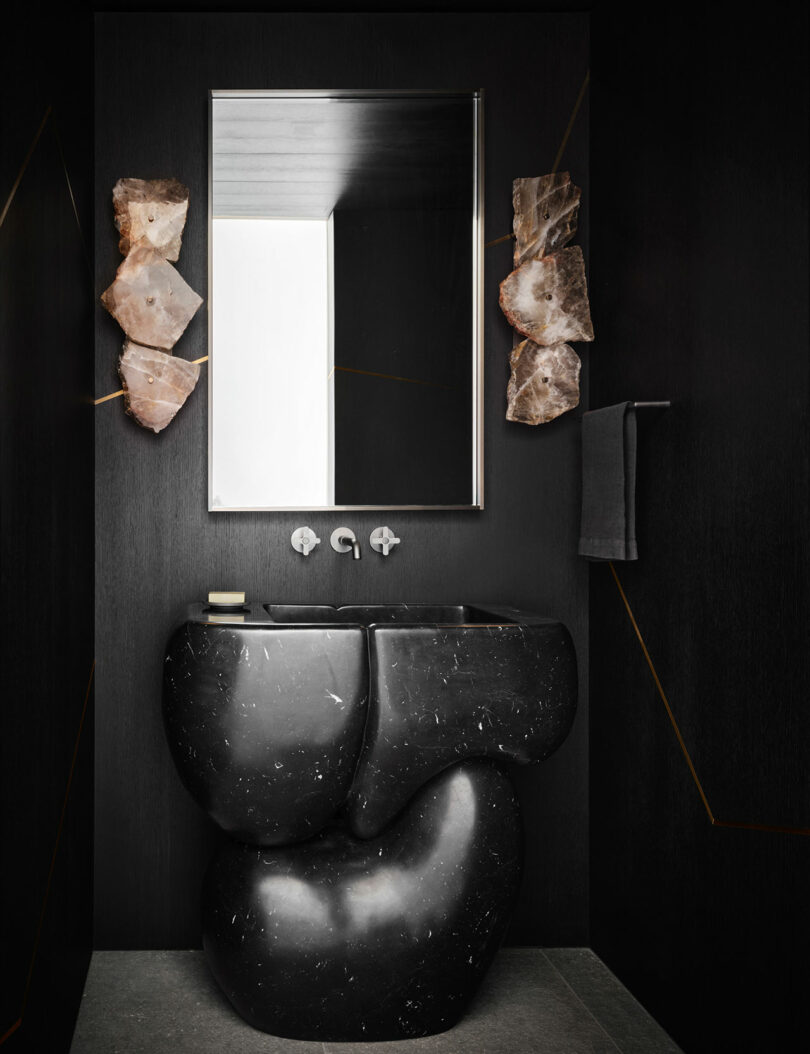 A modern black bathroom with an abstract sink, wall-mounted faucet, square mirror, and towel on a rack. Two rock-like light fixtures are on either side of the mirror.