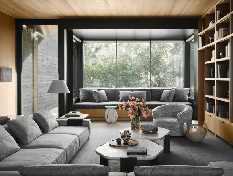 Modern living room with large windows, gray sofas, a curved chair, and a coffee table with flowers. Bookshelves line the wall, and greenery is visible outside.