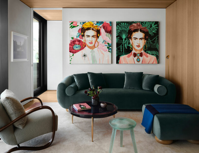 A modern living room with a green sofa, armchair, and a round coffee table. Two colorful portraits hang on the wall above the sofa. A window on the left provides natural light.