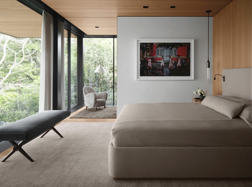 Modern bedroom with a large bed, a framed wall picture, and a sitting area by glass windows overlooking a green outdoor scene.