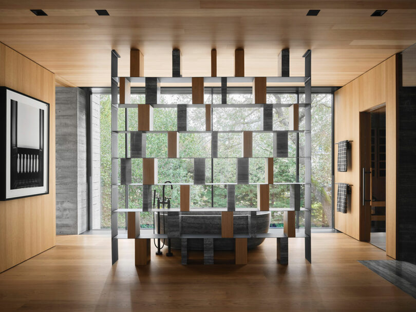 Modern bathroom with a freestanding tub and a geometric wooden shelf divider. Large windows provide a view of greenery outside. Light wood flooring and ceiling complement the minimalist design.