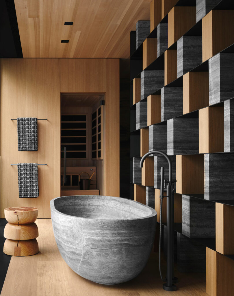 Modern bathroom with a stone bathtub, wooden flooring, and a geometric wall design. Two stacked wooden stools and patterned towels are visible.