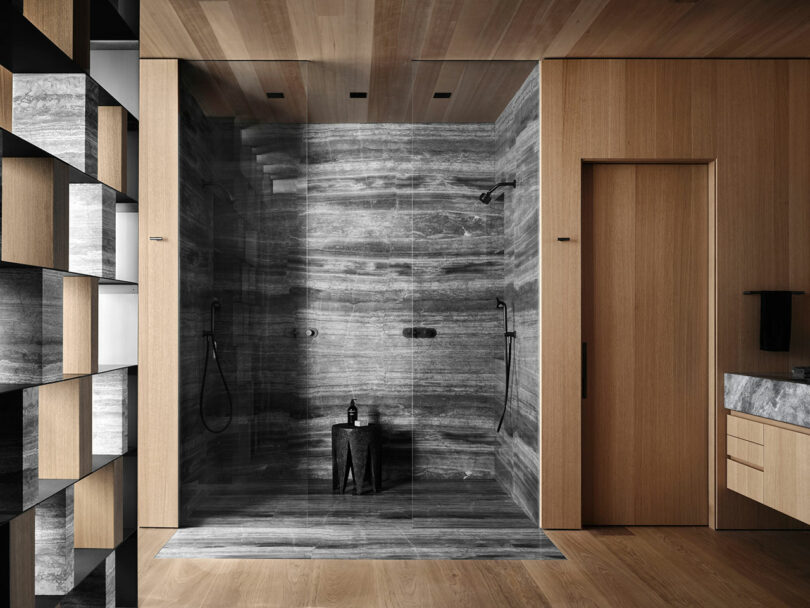 A modern bathroom features a large shower with gray stone walls, a black stool, and wooden accents. The room includes a wooden door and shelves.