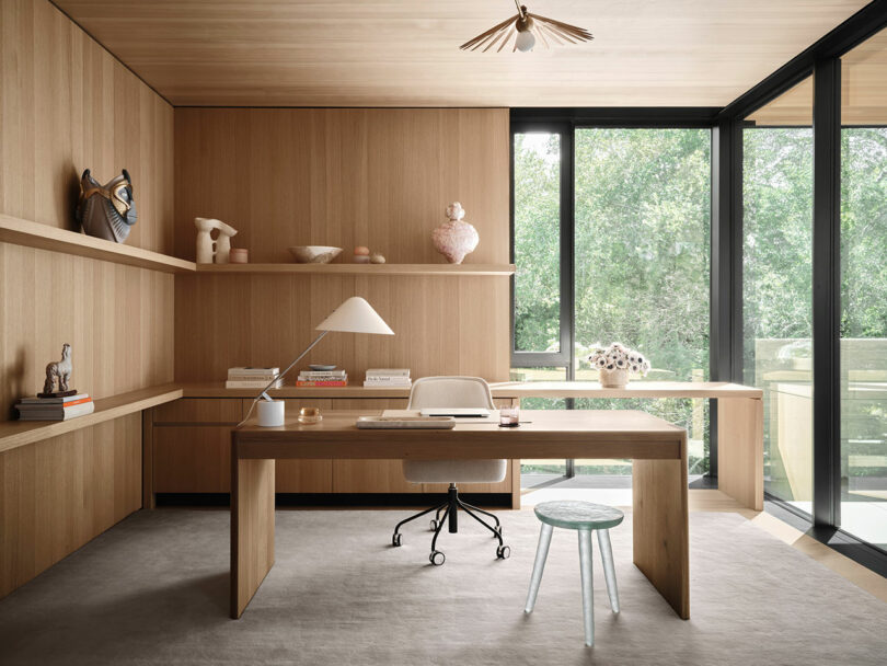 A modern home office with wooden walls and furniture, a desk with a lamp and chair, and large windows revealing a view of greenery outside.