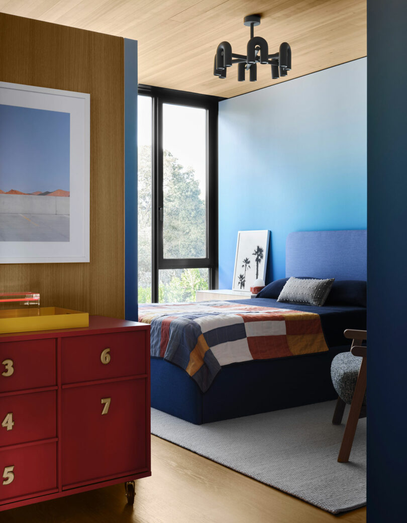 A modern bedroom with a blue gradient wall, bed with a multicolored quilt, a large window, red numbered drawers, and art on the floor. A black ceiling light fixture hangs overhead.