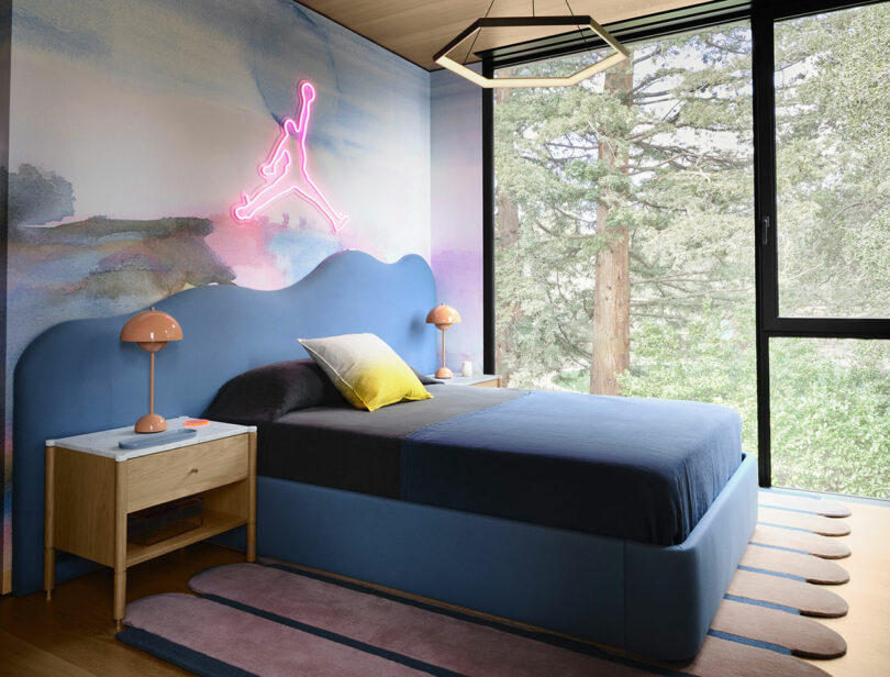 Modern bedroom with a blue bed, wavy headboard, two bedside tables with lamps, a neon sign on the wall, large window, and views of trees outside.